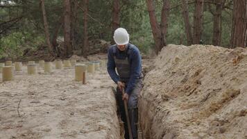 un trabajador vistiendo un blanco difícil sombrero es activamente excavación un estrecho zanja en un boscoso área. el rodeando terreno es desigual con visible árbol tocones y suelo muchísimo. video