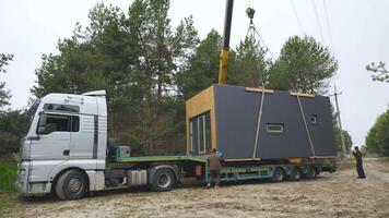 une grue est levage une préfabriqué maison de une bande annonce dans une forêt clairière pendant un de bonne heure Matin. ouvriers sont présent à guider le opération. video