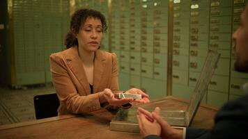 A woman in formal wear sits at a bank table, talking to a man video