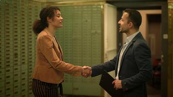 A man in a suit is seen writing on a clipboard in a bank vault, while a man and a woman talk to each other in the background. The plot revolves around a mysterious interaction in a highsecurity video