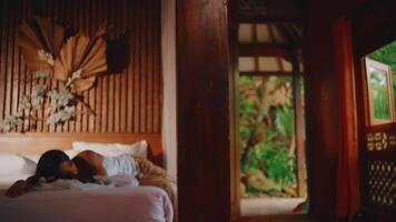 Cozy interior with a woman resting on a bed, warm lighting, and a view of greenery through an open window. video