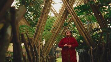 Person in red outfit standing contemplatively in a rustic wooden gazebo surrounded by greenery. video