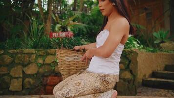 Serene woman in traditional attire holding a basket, sitting outdoors with lush greenery in the background. video