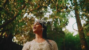 Elegant woman in a sparkling dress and tiara posing under a tree canopy with a serene expression. video