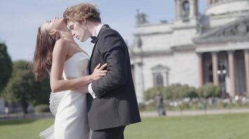 schön Jungvermählten. Aktion. ein Paar von Liebhaber, das Braut im ein fest Weiß Kleid und das Bräutigam im ein passen mit lange Haar Pose auf das Straße Nächster zu das Damm und schön historisch Gebäude im video