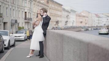 Beautiful newlyweds. Action. A couple of lovers, the bride in a tight white dress and the groom in a suit with long hair pose on the street next to the embankment and beautiful historical buildings in video