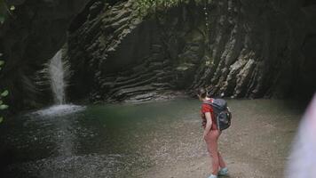 The girl looks at the waterfall. CREATIVE. Girl tourist looks at the waterfall in the mountains. Exotic place with a waterfall and a small lake in the mountains video