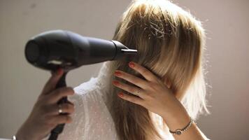 A young woman dries her hair with a hairdryer isolated on a beige wall background. Art. Young blond woman in a white shirt styling her hair. video