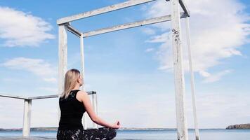 blond jung Frau Sitzung im Lotus Position in der Nähe von See und tun Atmung Übungen. Konzept. Mädchen im Sport passen Sitzung im Meditation auf ein Seebrücke durch das Wasser auf Blau wolkig Himmel Hintergrund. video