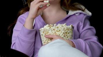 Young middle eastern lady spending her free time at home, watching TV and eating popcorn on sofa, free space. Excited lady with remote control enjoying interesting movie video