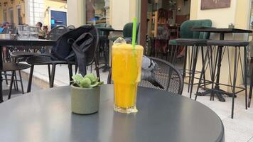 een duif vogel vliegt op de tafel in een cafe waar Daar is oranje sap met ijs, voedsel buiten, de sap is in een glas glas met een groen plastic rietje, een veel van ijs heeft stroomde op de tafel video