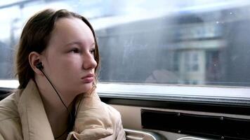 Tonårs flicka rider i skytrain på tåg i buss hörlurar i öron lyssnar till musik värma beige jacka hår är bunden ner verklig person sitter undangömt ben under ledsen drömmar väntar sluta i händer innehav telefon video