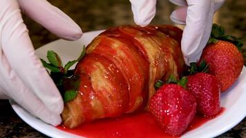 com luva mãos estão quebra delicioso fresco croissant encharcado dentro vermelho xarope instruções do delicioso sobremesa é jogando cintilante morangos estão em mesa Eu quer para comer Assistir pastelaria Verifica prontidão video