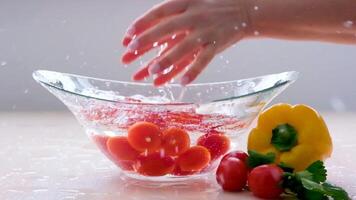 Fresh organic red cherry tomatoes with green tails fall into a glass transparent bowl with water with big splashing and spraying on a wooden table on the background of a white brick wall. Slow motion. video