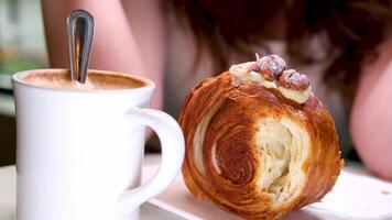 Frau halt gebissen Brötchen, Aussicht durch Glas von Cafe Zimmer, mit Betrachtung. Besucher Gesicht nicht im rahmen, sie halt Smartphone im richtig Hand, Art Botschaft. Tasse von Kaffee auf das Tabelle video