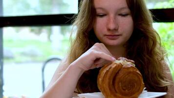 a young girl eats a round croissant in a restaurant by the window, she picks up cream with her little finger and licks the delicious cream from her finger, a delicacy to taste adolescence video