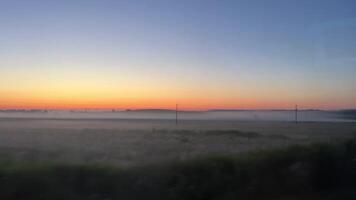 Visualizações a partir de a ônibus janela em a campo dentro a névoa pôr do sol alvorecer piscando árvores azul céu vermelho horizonte névoa video