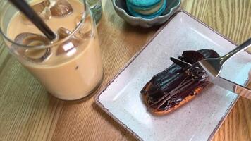 ein Stück von Pudding Kuchen bedeckt mit dunkel Schokolade ist Schnitt aus mit ein Messer und Gabel in der Nähe von ein Glas von vereist Kaffee das Teller zappelt auf ein hölzern Tabelle es ist ungünstig zu Schnitt video