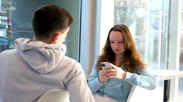 liv av tonåringar på de telefon en pojke och flicka sitta på tabell förbi fönster varje med deras smartphone spendera tid på internet i social nät Kafé skola cafeteria ta bilder av varje Övrig skjuta videoklipp video