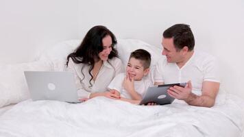 husband wife and son laugh a lot funny joke bed holiday look at tablet communicate conversation love at home happiness joy success in gadgets white background place for advertising text happy family video