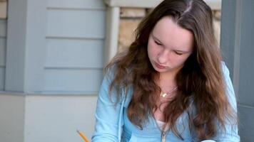 teenager girl does homework on porch of house she writes with pencil in large notebook notebook in folder next to backpack autumn summer sit on threshold beautiful young woman writes draws solves video
