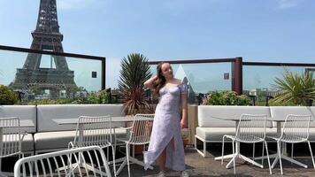 ein schön Teenager Mädchen im ein lange Blau Kleid steht gegen das Hintergrund von das Eiffel Turm im ein Restaurant, sie richtet sich auf ihr Haar und Blätter das Kleid ist geblasen durch das Wind von Raum zum Text video