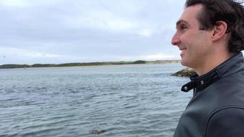 une Beau français homme dans noir veste des stands sur le rivages de le atlantique océan, il a noir cheveux, une cuir veste et il mouches avec le sien mains à le côtés Voyage La publicité video