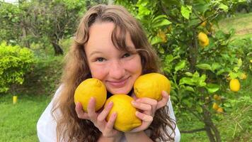 un Adolescente niña en un blanco blusa con rubio pelo soportes en contra el antecedentes de un limón árbol ella sostiene Tres desplumado limones en su manos ella es feliz, satisfecho y hace un Beso con su labios video