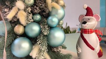 A little mouse in a New Year's hat with a bag and a scarf stands under tree Christmas background New Year Green Christmas tree with white snow on it blue and silver balls Nice pastel light color video