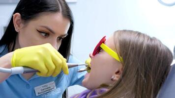 dentale Riempimento nel bambini latte denti deteriorare velocemente dentista colpi aria su un' carie buco e sembra attraverso esso con un' specchio ragazza si siede nel un' dentale sedia nel occhiali da sole giovane bellissimo vero persone video