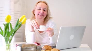 mujer a ventana Bebiendo té café cerca computadora chateando con amigos gasto agradable hora relajante trabajando como persona de libre dedicación ganador dinero fácilmente teniendo divertido y relajante desayuno en mesa flores video