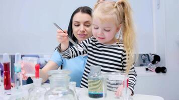 a little fair-haired girl with ponytails on her head and a striped blouse takes out pill from flask with tweezers and adds chemical experiments to a glass bottle with blue paint. children's laboratory video