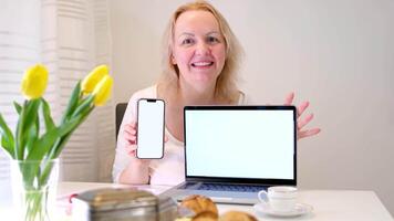 lindo meia idade mulher apontando dela dedo às a telefone e computador portátil monitor com branco fundo para publicidade texto em a mesa café da manhã copo do café amarelo tulipas acolhedor casa branco paredes video