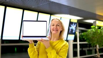 schön mittleren Alters Frau beim das Flughafen steht im ihr Hände mit ein Laptop Werbung ein Weiß Monitor Raum zum Text lächelnd zeigt an Tanzen hell Kleider Anzeigetafel video