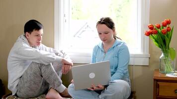 girl and boy teenagers doing homework or watching movies on laptop sitting on small sofa against window boy bit lip looking intently girl shows smiling at monitor screen real people life study video