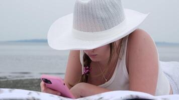 bonito rubio mujer tendido abajo mientras ella es leyendo un libro electronico niña en playa con móvil teléfono, blanco sombrero acostado en albañiles acecho video