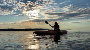 adolescent fille est kayak à le coucher du soleil dans pacifique océan, seulement le silhouette de kayak pagaies est visible elle nage le long de ensoleillé chemin à le mer. une magnifique ciel avec petit des nuages et le Soleil brille par video