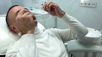 bonito adulto homem desgasta transparente óculos dentro a dental cadeira limpeza dele dentes ele é esperando para a médico branco roupas branco travesseiro e todos a Mais recentes tecnologias por aí profissional clínica video