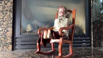 An old and very strange doll sits in a rocking chair against the background of a fireplace, it sways very scary, as if in a horror movie, as if something should happen or has already happened video