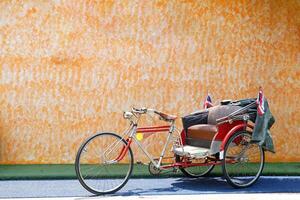 An old tricycle stops as a check-in point at a restaurant with an orange cement wall background. In the past, it was a vehicle that Thai people preferred to use. Currently serving tourists. photo