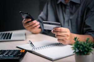 hombre con crédito tarjeta y teléfono inteligente finaliza un transacción, con un calculadora y bloc cercano. en línea pago transacción mi comercio compras Internet Finanzas bancario negocio concepto. foto