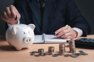 Person in suit depositing coin into piggy bank on desk with coins and calculator. investment and save money tax finance management concept photo