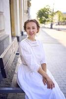 elegant middle age woman in a white vintage dress sitting on a bench in the morning city photo