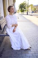 elegante medio años mujer en un blanco Clásico vestir sentado en un banco en el Mañana ciudad foto