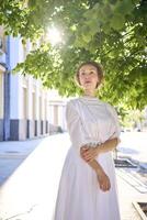 elegante medio años mujer en un blanco Clásico vestir en contra el antecedentes de histórico edificios en el Mañana ligero foto