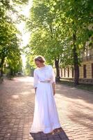 elegante medio años mujer en un blanco Clásico vestir en un iluminado por el sol callejón foto