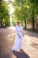 elegante medio años mujer en un blanco Clásico vestir en un iluminado por el sol callejón foto