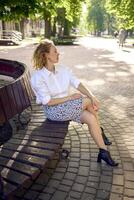 beautiful middle age woman in 70s, 80s style clothes on a bench in a sunlit avenue photo