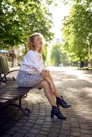 beautiful middle age woman in 70s, 80s style clothes on a bench in a sunlit avenue photo