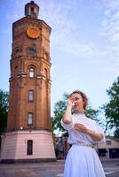 elegante joven mujer en un blanco Clásico vestir en el cuadrado cerca el histórico agua torre en vinnytsia foto